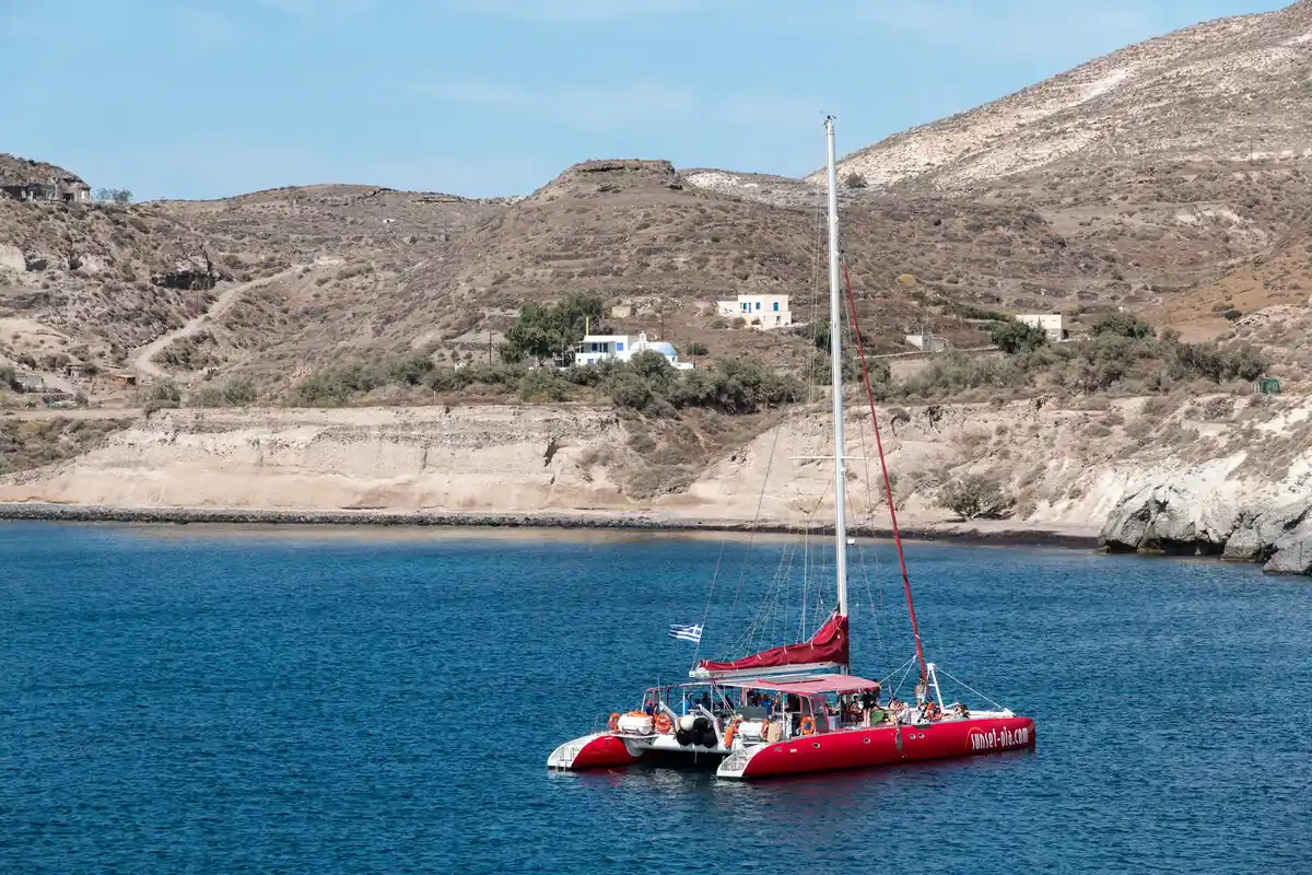 Red sailing catamaran with tour passengers anchored in a bay below the cliffs of Akrotiri, Santorini