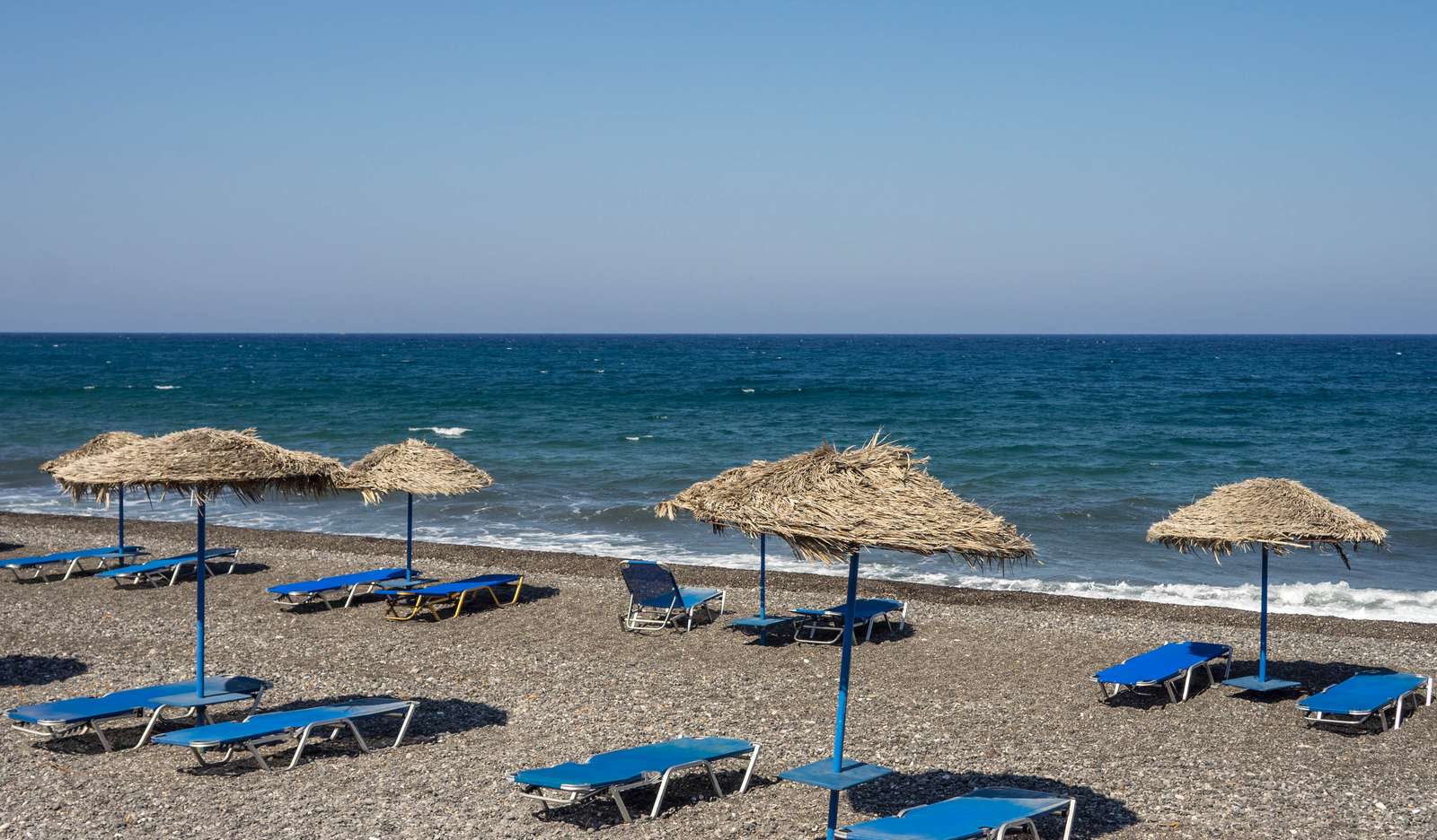 Strohschirme und blaue Liegen auf dem dunklen Vulkansand des Strands von Kamari, Santorini