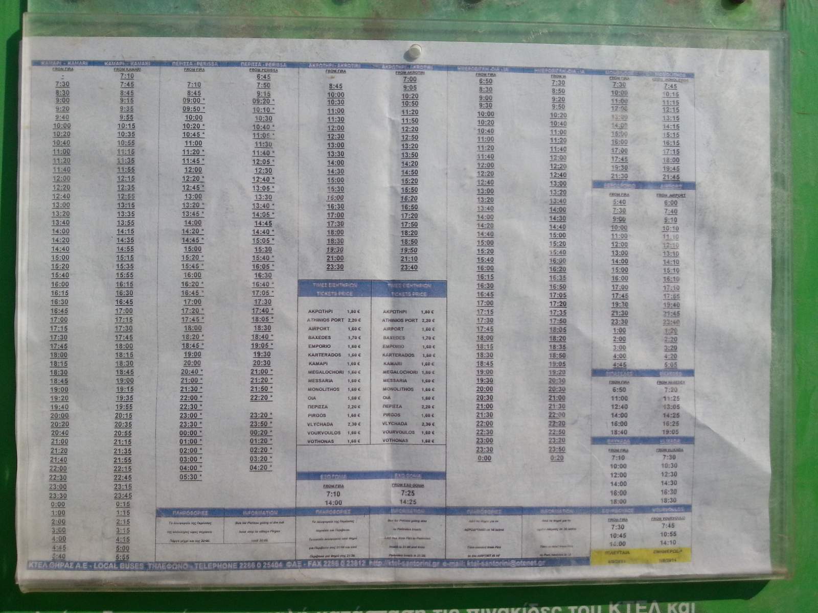 Printed KTEL timetable board behind plastic at a Santorini bus shelter, with departure columns per destination and a ticket-price list